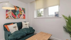 Una sala de estar con un sofá color turquesa, una mesa de centro de madera, un cuadro abstracto de colores y una ventana con persiana blanca.