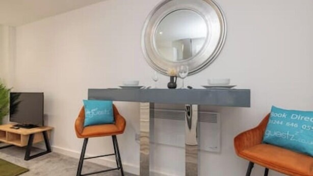 Una sala de estar moderna con un televisor, dos taburetes altos, un espejo redondo y una mesita con un florero y un vaso.