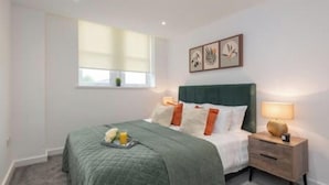 Un dormitorio con una colcha verde, ropa de cama blanca y una mesita de noche con lámpara.
