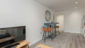 Een moderne woonkamer met een flatscreen televisie, een houten salontafel en een eethoek met twee barkrukken.