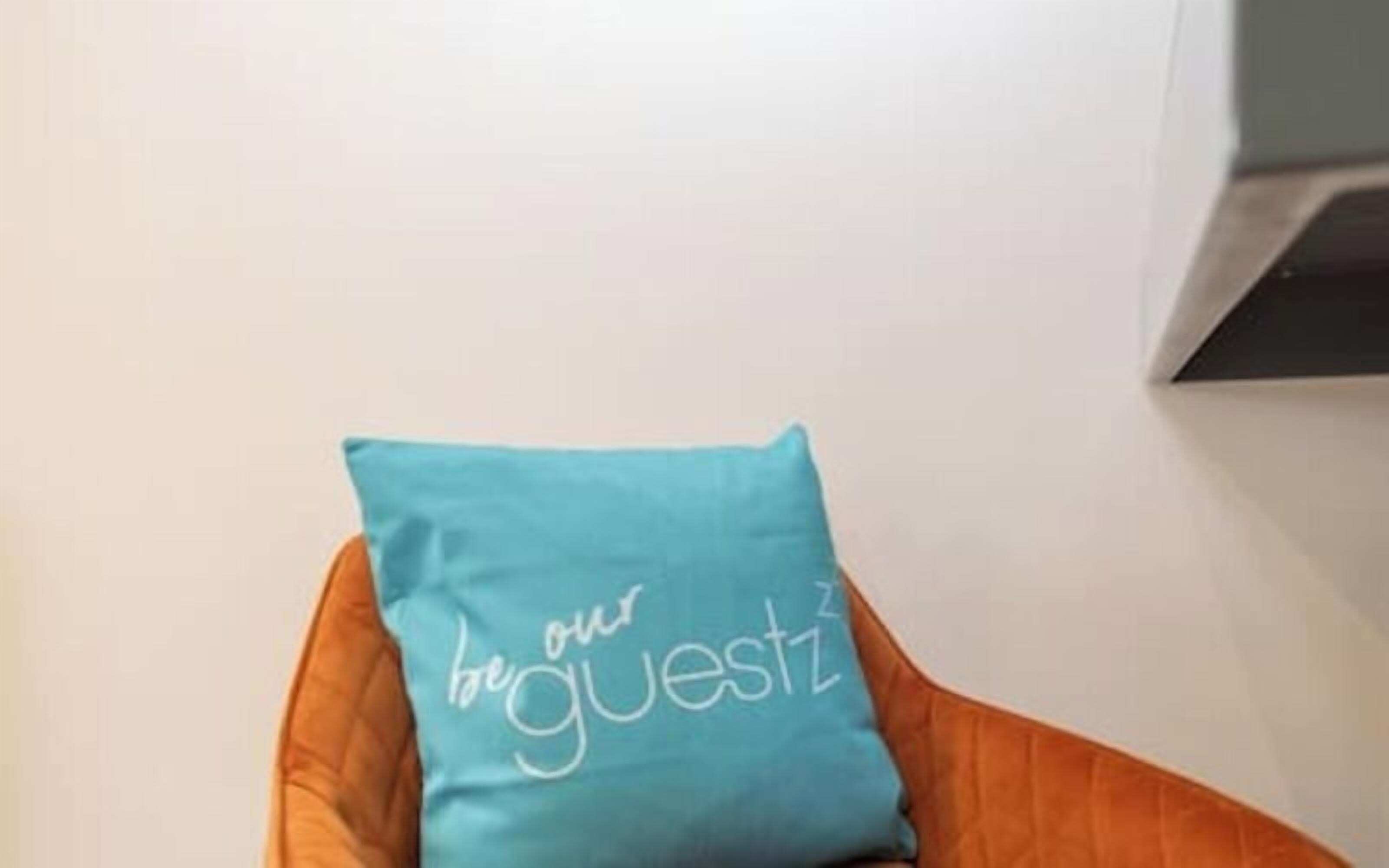 A teal pillow with the text 'be our guest' on a chair.