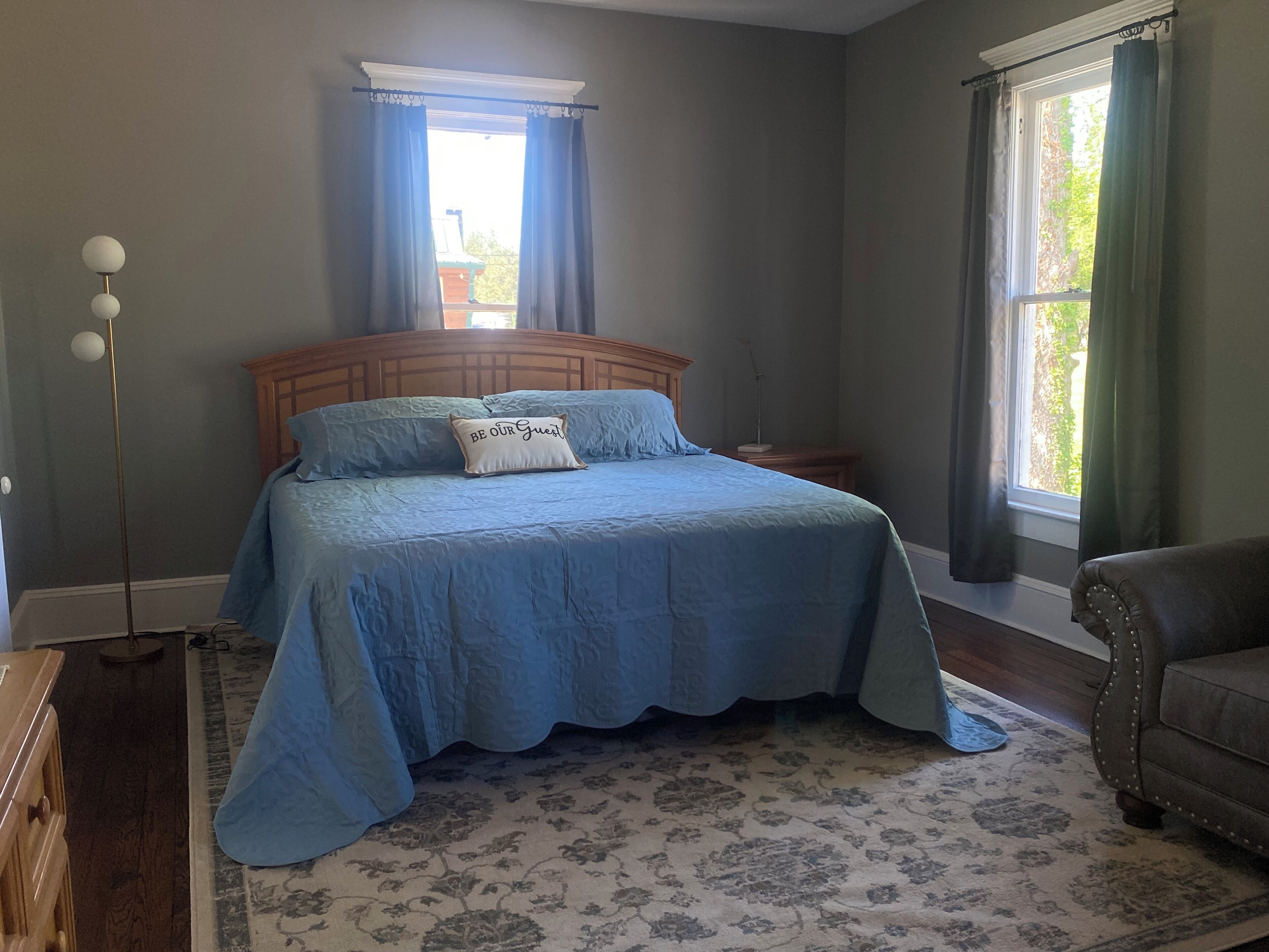 Quarto com cama grande, mesa de cabeceira, luminária de chão e vista para o exterior pela janela.