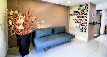 Un moderno vestíbulo de hotel con un sofá gris, una planta decorativa y una pared con saludos en varios idiomas.