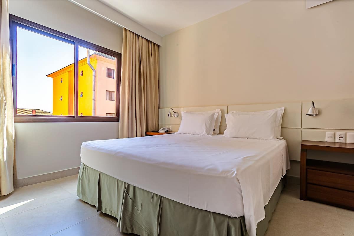 Une chambre d’hôtel avec un grand lit, une table de chevet, une commode, une fenêtre avec des rideaux et une vue sur les bâtiments environnants.
