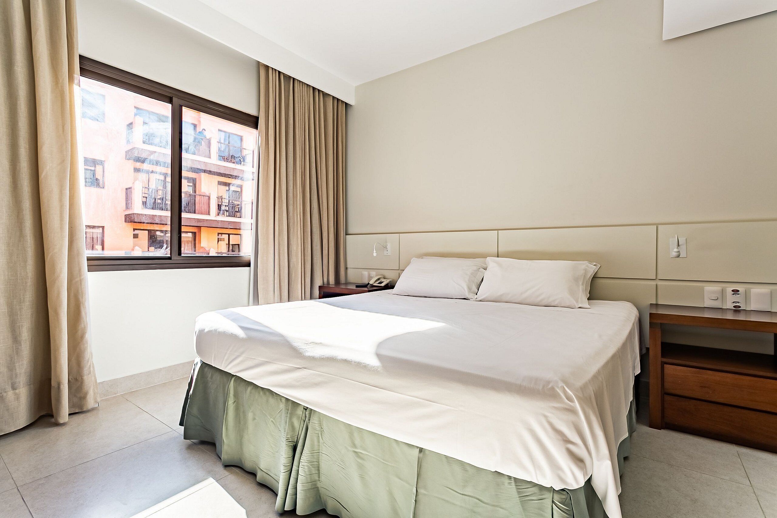 A hotel room with a bed, a nightstand, a window with curtains, and a view of buildings outside.