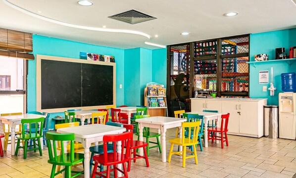 Un aula con sillas y mesas coloridas, una pizarra y material educativo en estanterías.