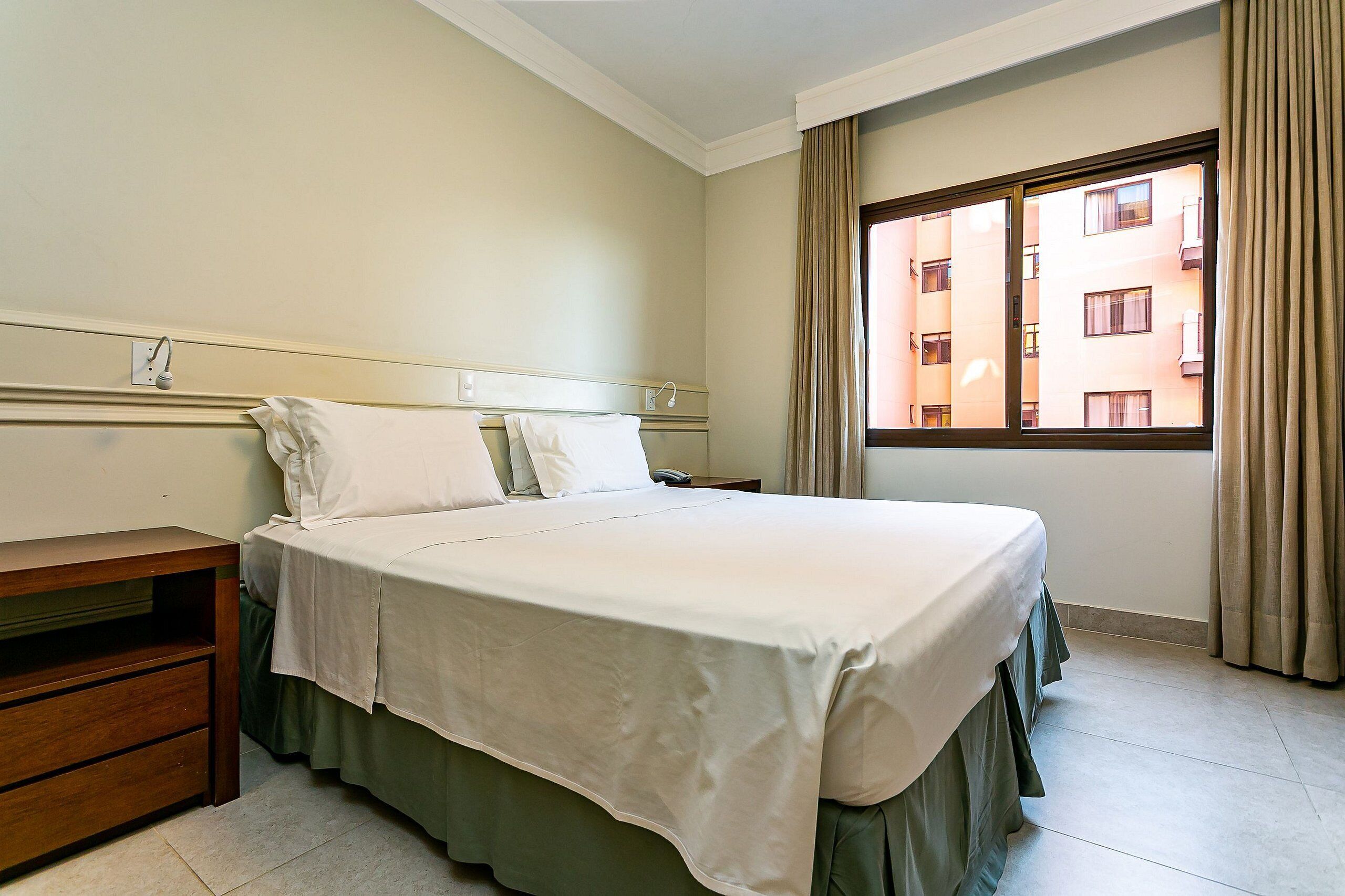 A hotel room with a bed, bedside table, and a window with curtains.