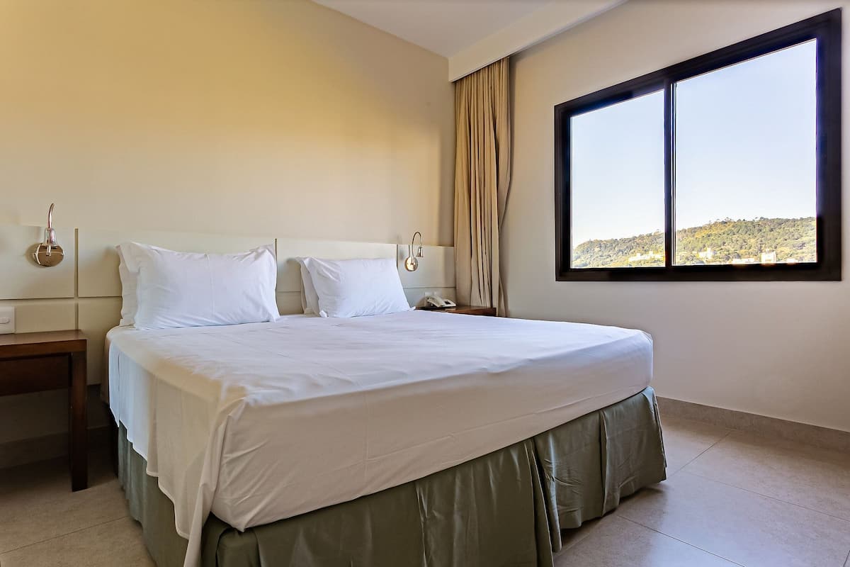 Une chambre d’hôtel avec un grand lit, une table de chevet, une commode, une fenêtre donnant sur un beau paysage et un rideau.