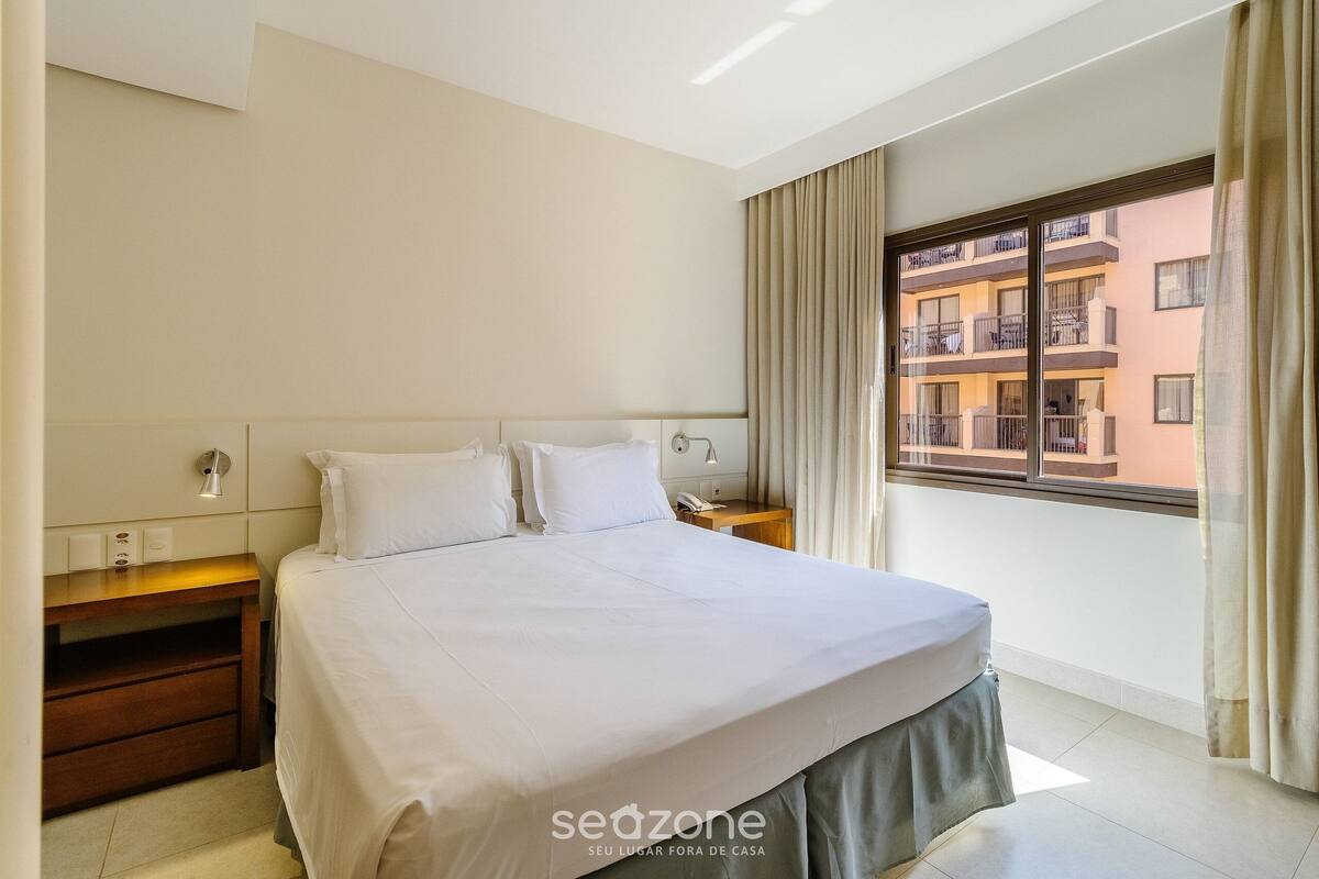 A hotel room with a bed, bedside tables, a window with curtains, and a view of buildings outside.