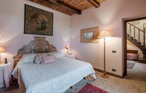A bedroom with a large bed, a wooden headboard, a painting above the bed, a lamp, and a staircase in the background.