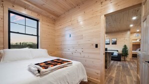 A wooden cabin room with a bed, a window with a view, a small tree, and a door leading to another room. - Cozy Trailside Lodge #2 (Boonville)