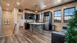 A modern kitchen with wooden walls, a central island, stainless steel appliances, and large windows. - Cozy Trailside Lodge #2 (Boonville)