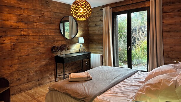 A bedroom with a wooden wall, a round mirror, a bedside table with a lamp, a bed with a beige blanket, and a window with curtains. - Chalet de l'Ailly (Varengeville-sur-Mer)
