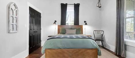 Un dormitorio con cama, una silla, una ventana con cortinas y una puerta.
