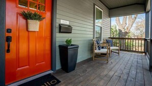 Une porte rouge avec une pancarte « Bienvenue », une boîte aux lettres noire, un panier en osier rempli de verdure, un pot de fleurs noir et une chaise en bois avec un coussin, le tout sur une terrasse en bois.