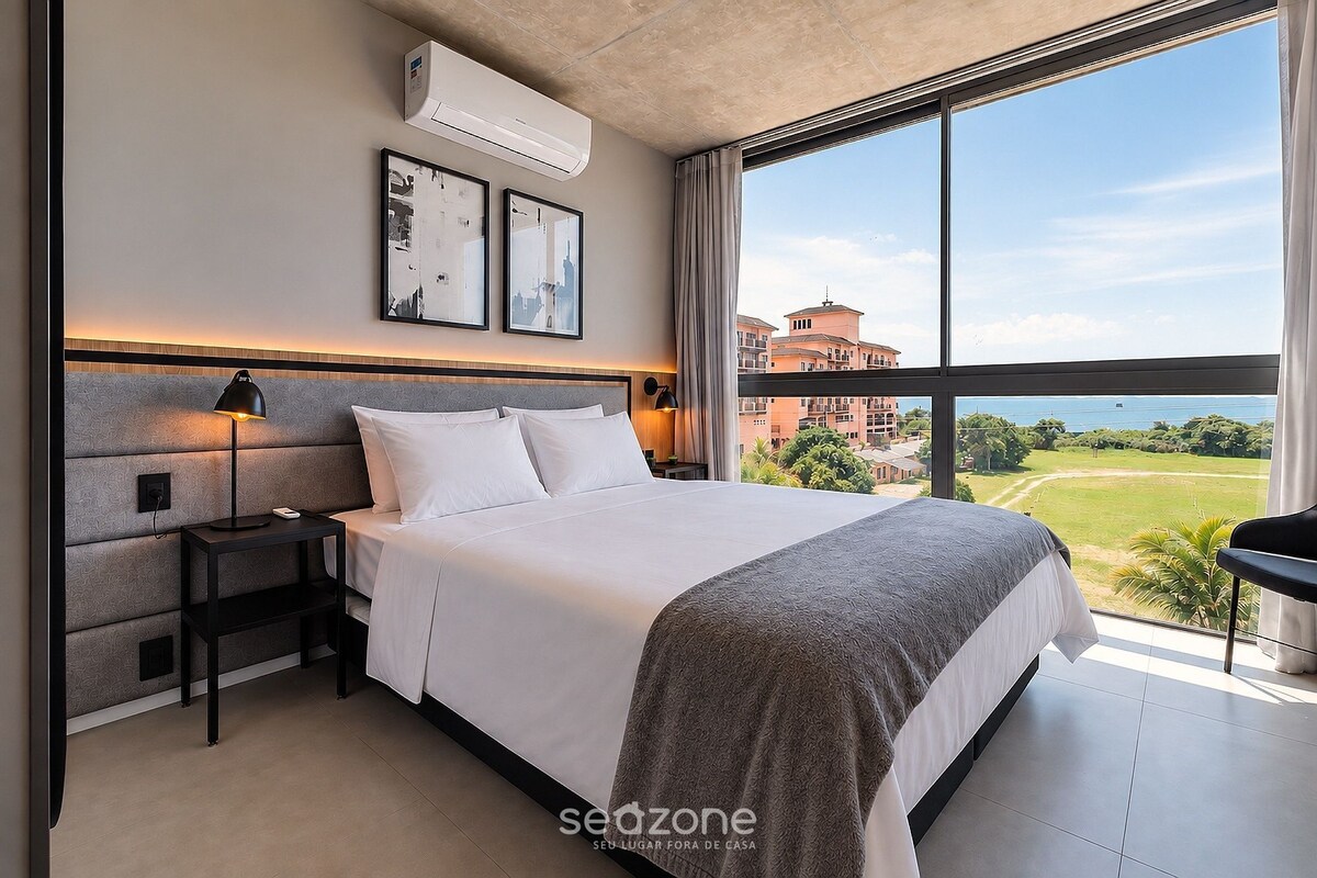 A modern hotel room with a large bed, a nightstand, a desk, and a view of a building and landscape through large windows.