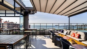 A rooftop terrace with tables and chairs, a sofa with cushions, and a view of the sea and buildings. - Spot Jurere sofisticação beira mar SPJs (Florianópolis)