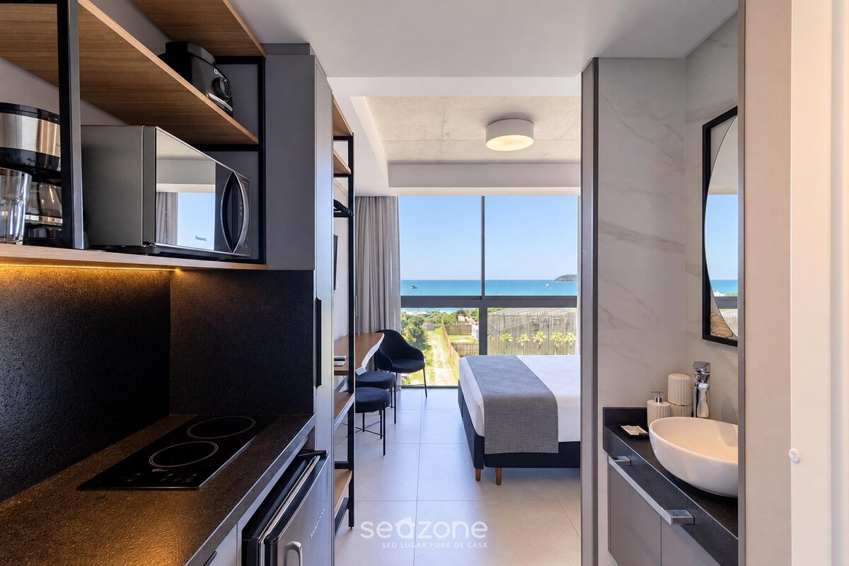 A modern hotel room with a kitchenette, a large bed, a bathroom with a sink, and a view of the sea.