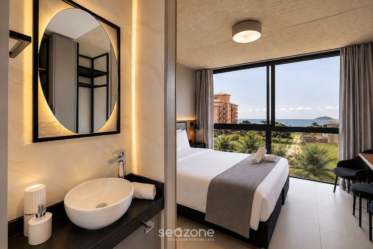 A hotel room with a large bed, a round mirror, a sink, and a view of the sea.