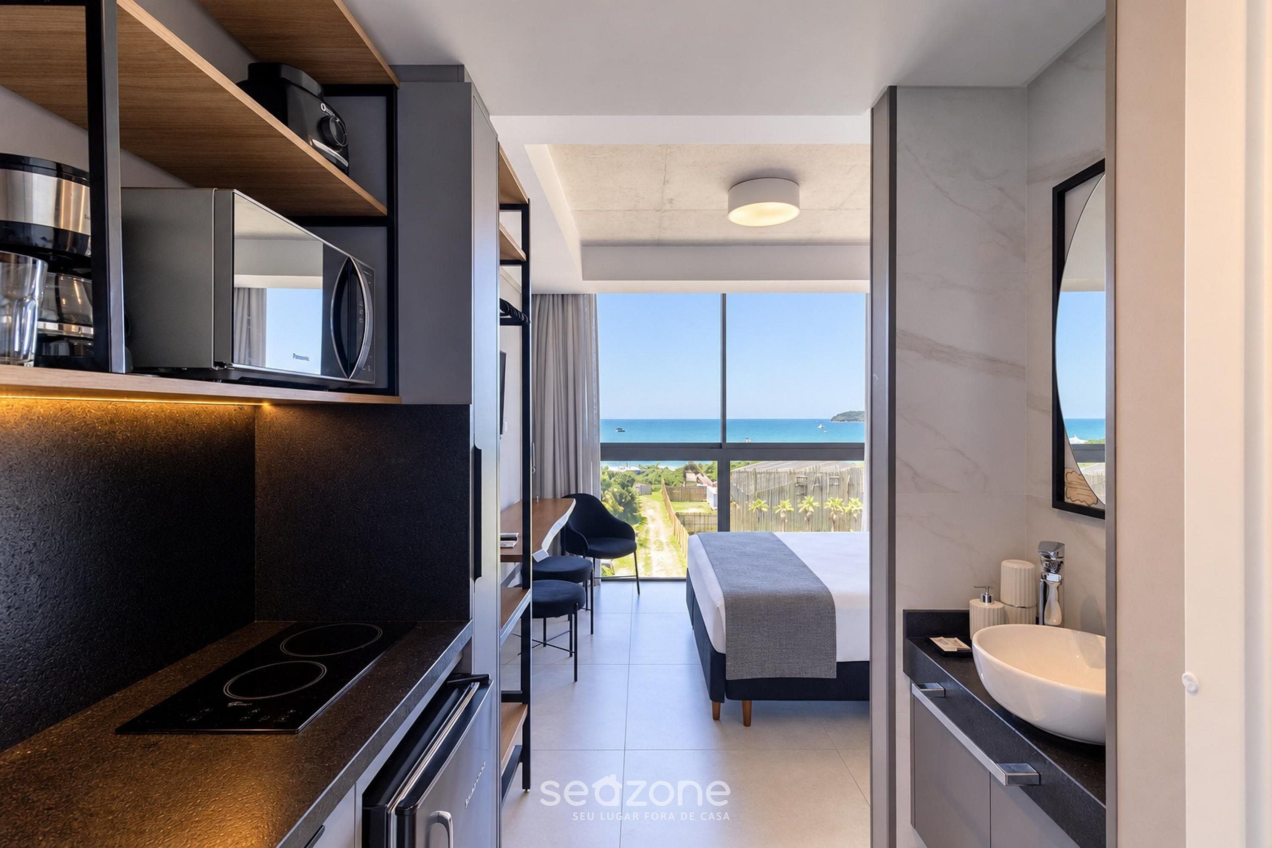 A modern hotel room with a kitchenette, a large bed, a bathroom with a sink, and a view of the sea.