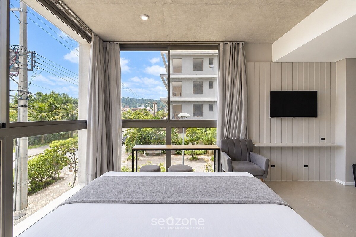A modern hotel room with a large bed, a TV mounted on the wall, and a balcony with a view of the surrounding area.