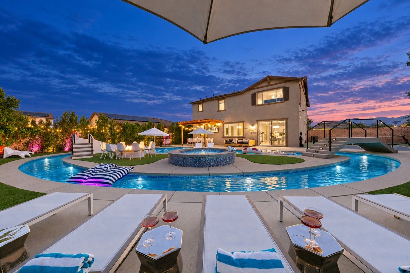 Una piscina con sillones de descanso y sombrillas, un porche cubierto y una casa con fachada iluminada.