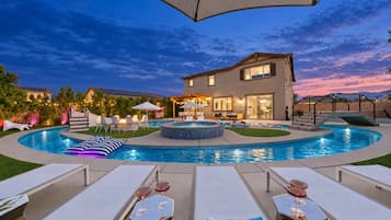 Une piscine avec des chaises longues et des parasols, un patio couvert et une maison dont l’extérieur est bien éclairé.