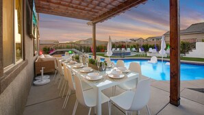 A covered outdoor dining area with a pool view, white chairs, and a table set for a meal. - Lazy River Pool+Spa+Pickleball+Game Room+BBQ+Golf (Coachella)