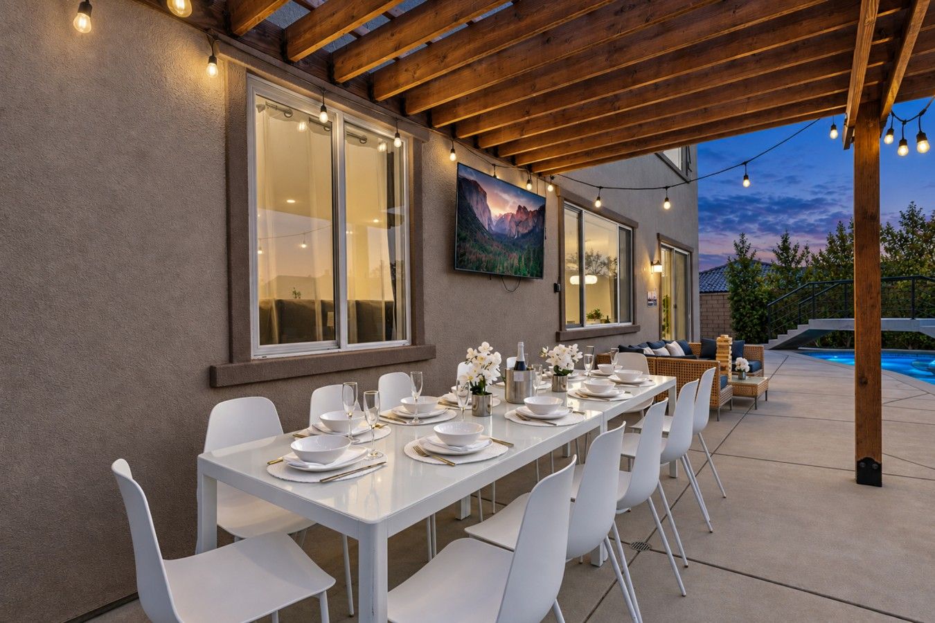 Un espace de restauration en plein air avec une longue table dressée pour un repas, entourée de chaises blanches, sous une pergola en bois illuminée de guirlandes lumineuses.