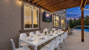 Un espace de restauration en plein air avec une longue table dressée pour un repas, entourée de chaises blanches, sous une pergola en bois illuminée de guirlandes lumineuses.