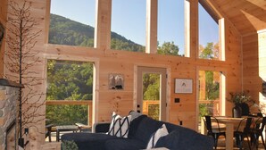 Eine Holzhütte mit Kamin aus Stein, einer Couch, einem Esstisch mit Stühlen und großen Fenstern mit Blick auf den Wald.