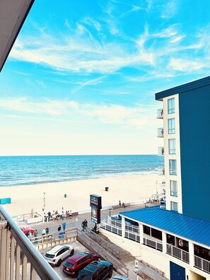 A beachfront hotel with a view of the ocean, a parking area with several cars, and a boardwalk with people walking.