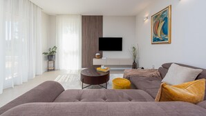 A modern living room with a large grey sofa, a round coffee table, and a flat-screen TV mounted on the wall. There is a painting on the wall and a potted plant on a side table. - Beach front home in Sukosan (Sukosan)