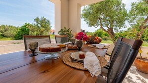 A wooden table set with a cake, pastries, and a fruit bowl, surrounded by chairs and a vase of flowers, with a view of trees and a road. - Beach front home in Sukosan (Sukosan)