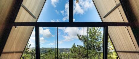 Ein Zimmer mit großem Fenster, einer Holzterrasse und Blick auf Bäume und Hügel.