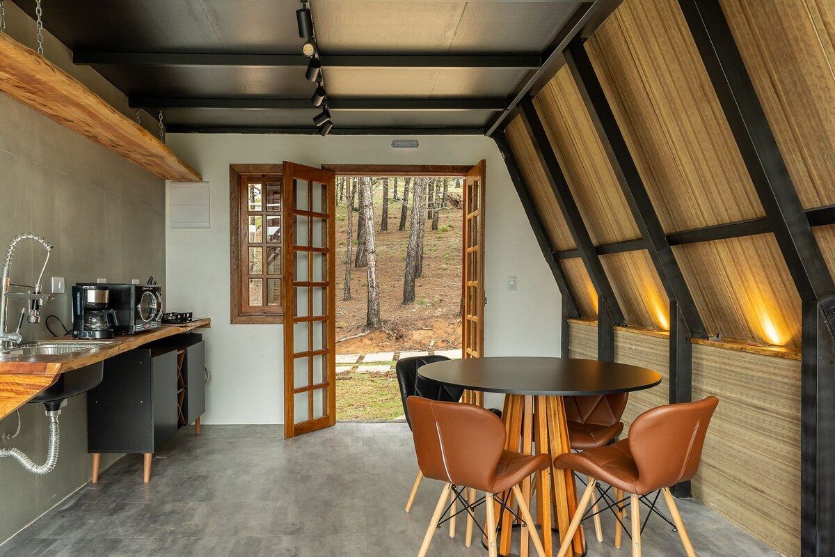 A modern kitchen with a wooden countertop, a coffee machine, and a sink. There is a dining area with a round table and two chairs. The room has a sloped ceiling and a large window with a view of trees.