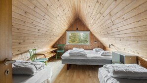 Um quarto de sótão aconchegante com paredes de madeira, uma janela grande, duas camas, uma cadeira e uma mesinha.