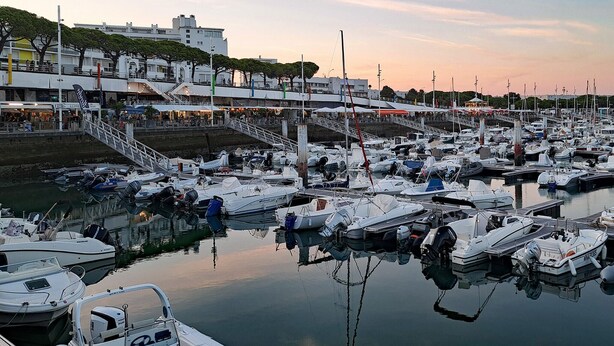 Un port de plaisance où sont amarrés de nombreux bateaux, un pont piétonnier et des bâtiments en arrière-plan.
