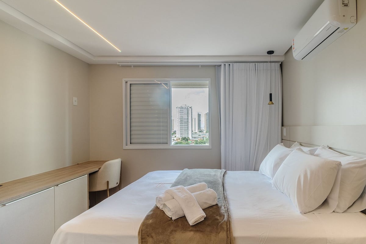 A hotel room with a bed, a desk, a chair, a window with curtains, and an air conditioning unit.