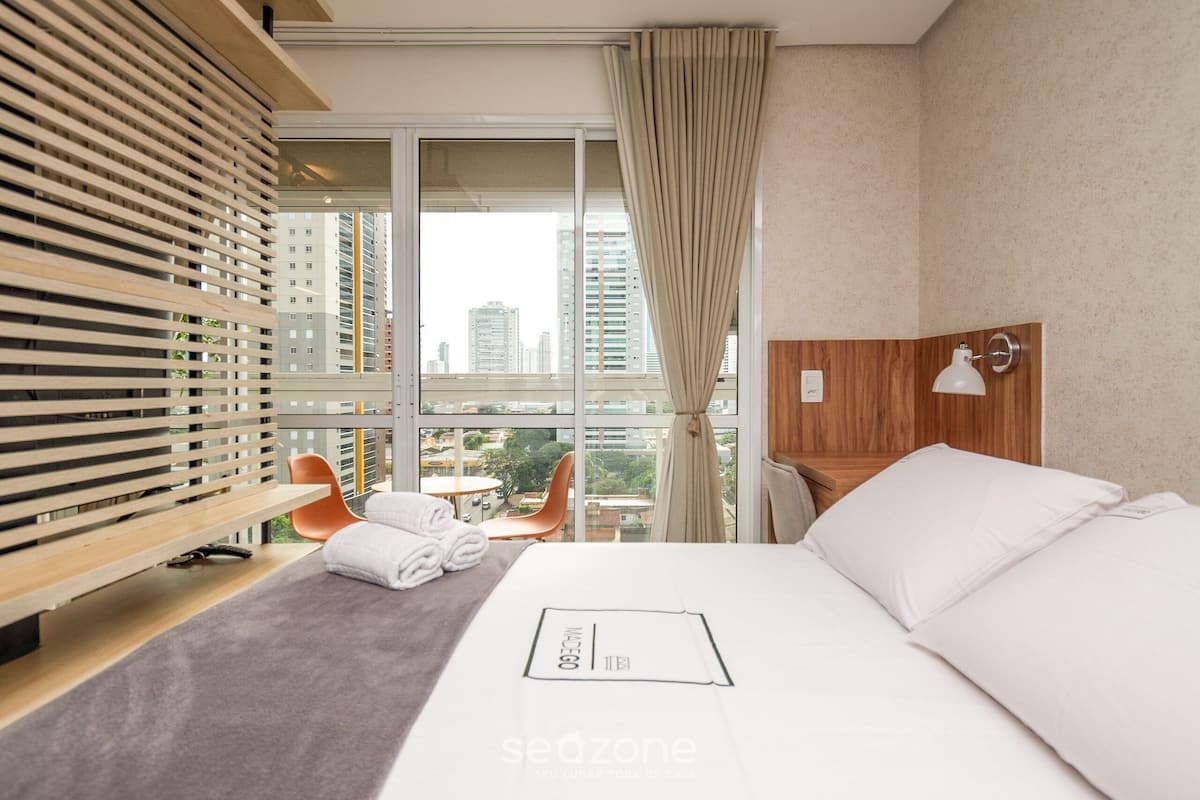 A hotel room with a large window, a bed, a wooden headboard, a chair, and a small table.