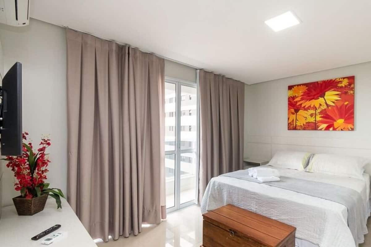A hotel room with a bed, a desk with a remote control, a potted plant, and a window with curtains.