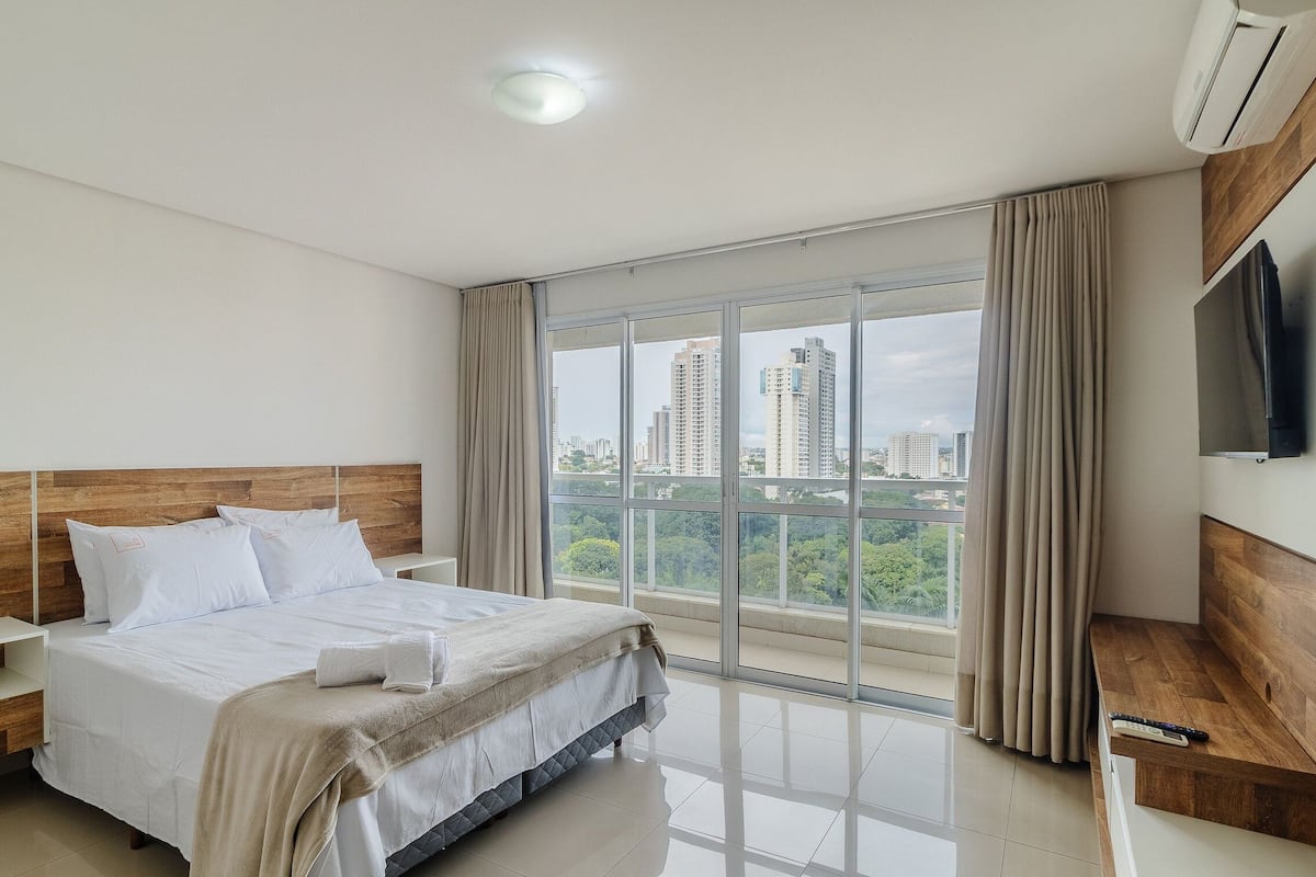 A modern hotel room with a large bed, a TV, and a view of tall buildings.