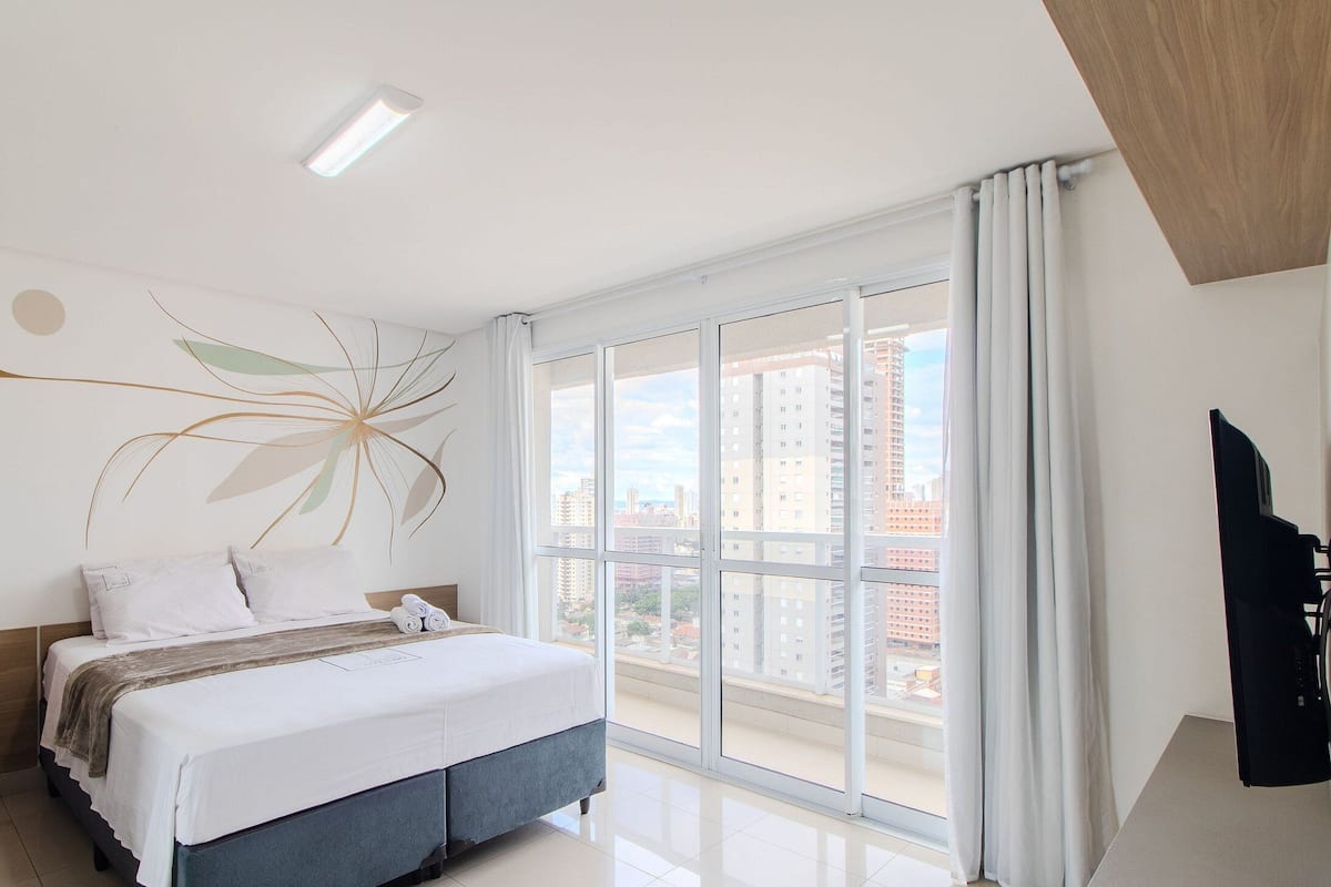 A modern bedroom with a large bed, a view of buildings through the window, and a flat-screen TV on the right.