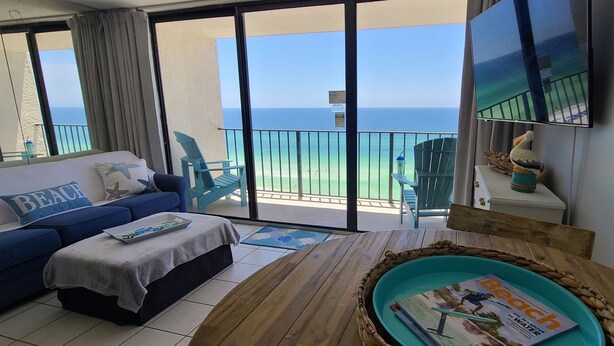 A balcony with a view of the ocean, a blue sofa with beach-themed pillows, a wooden table with a magazine, and a wicker tray. - Beachfront  Private Balcony 
 Edgewater Beach Resort
 Best Gulf Views (Panama City Beach)