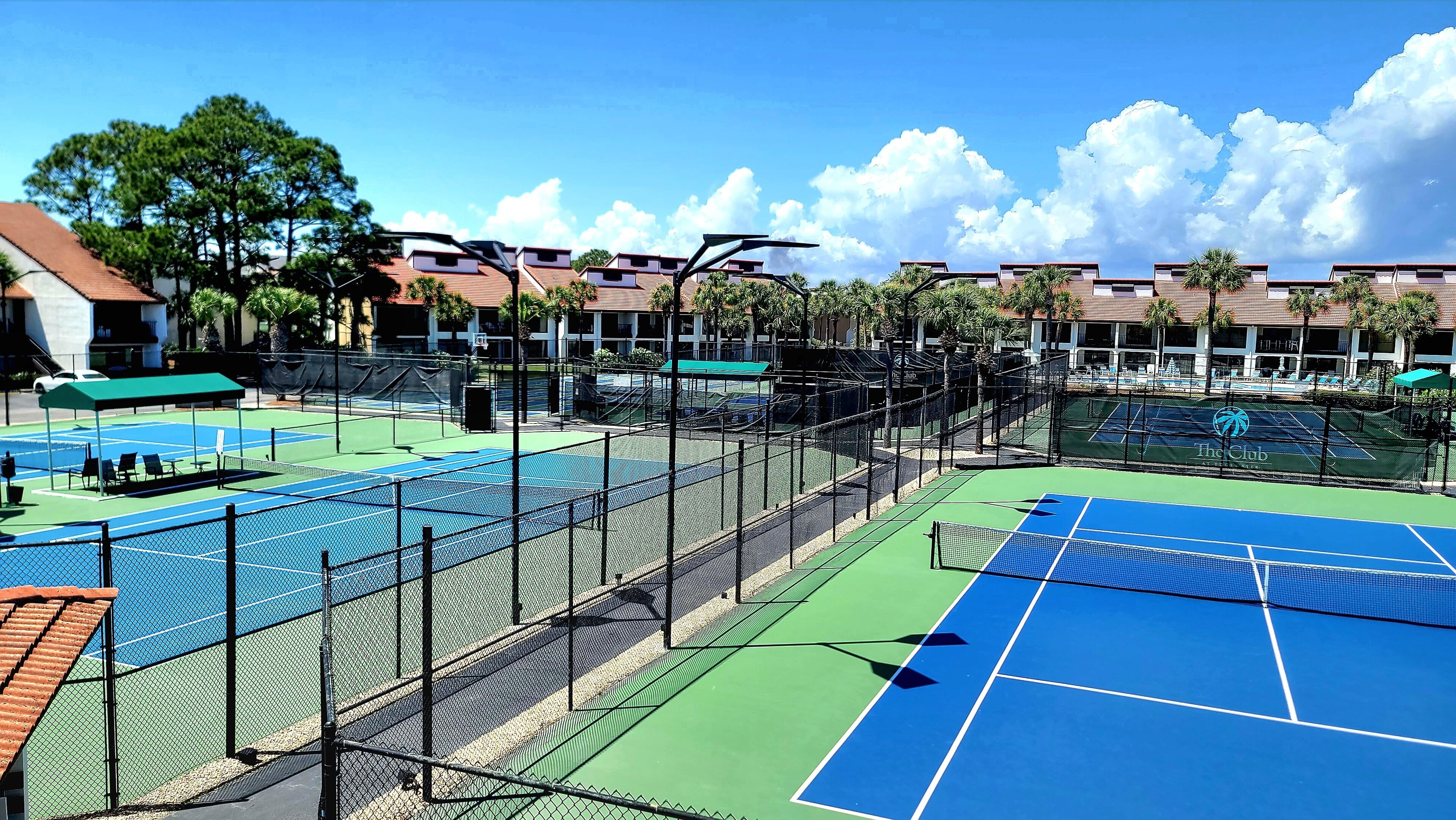 Una cancha de tenis con una red, rodeada por una cerca, y junto a una zona de piscina con sillas de descanso.