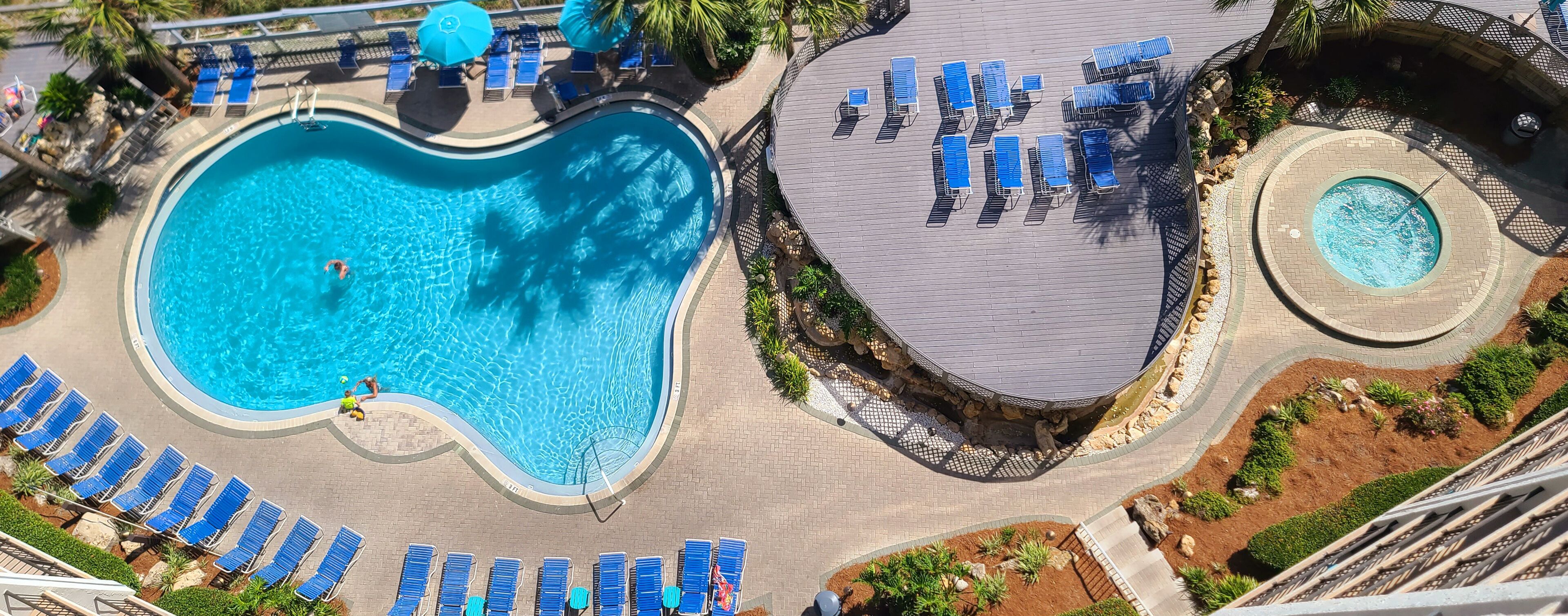 Una piscina grande con tumbonas y una piscina circular más pequeña, rodeada de zonas ajardinadas y un área de descanso cubierta.