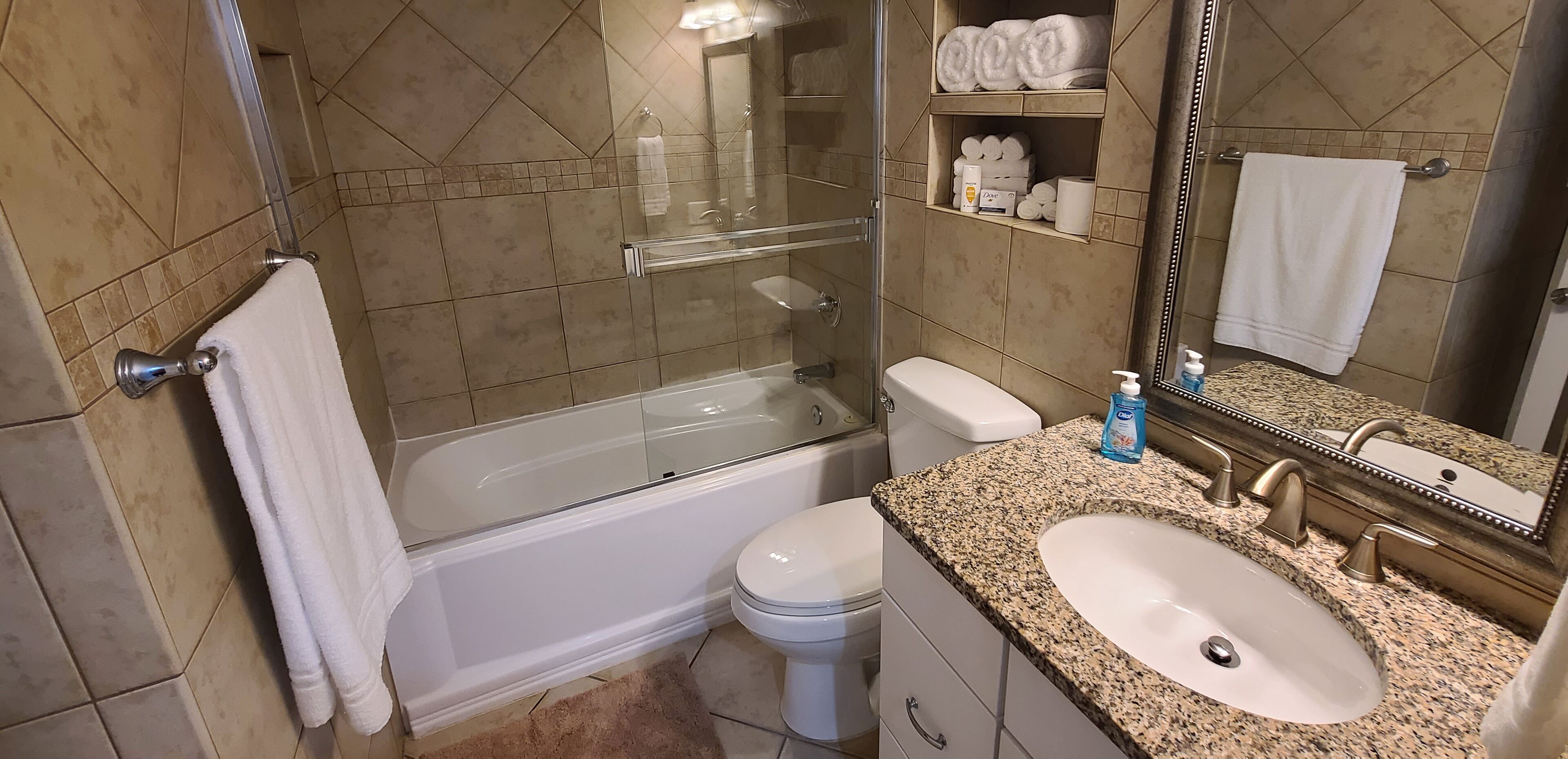 Un baño con ducha envuelta en vidrio, un inodoro blanco, un lavamanos con encimera de granito y un espejo con toallero.