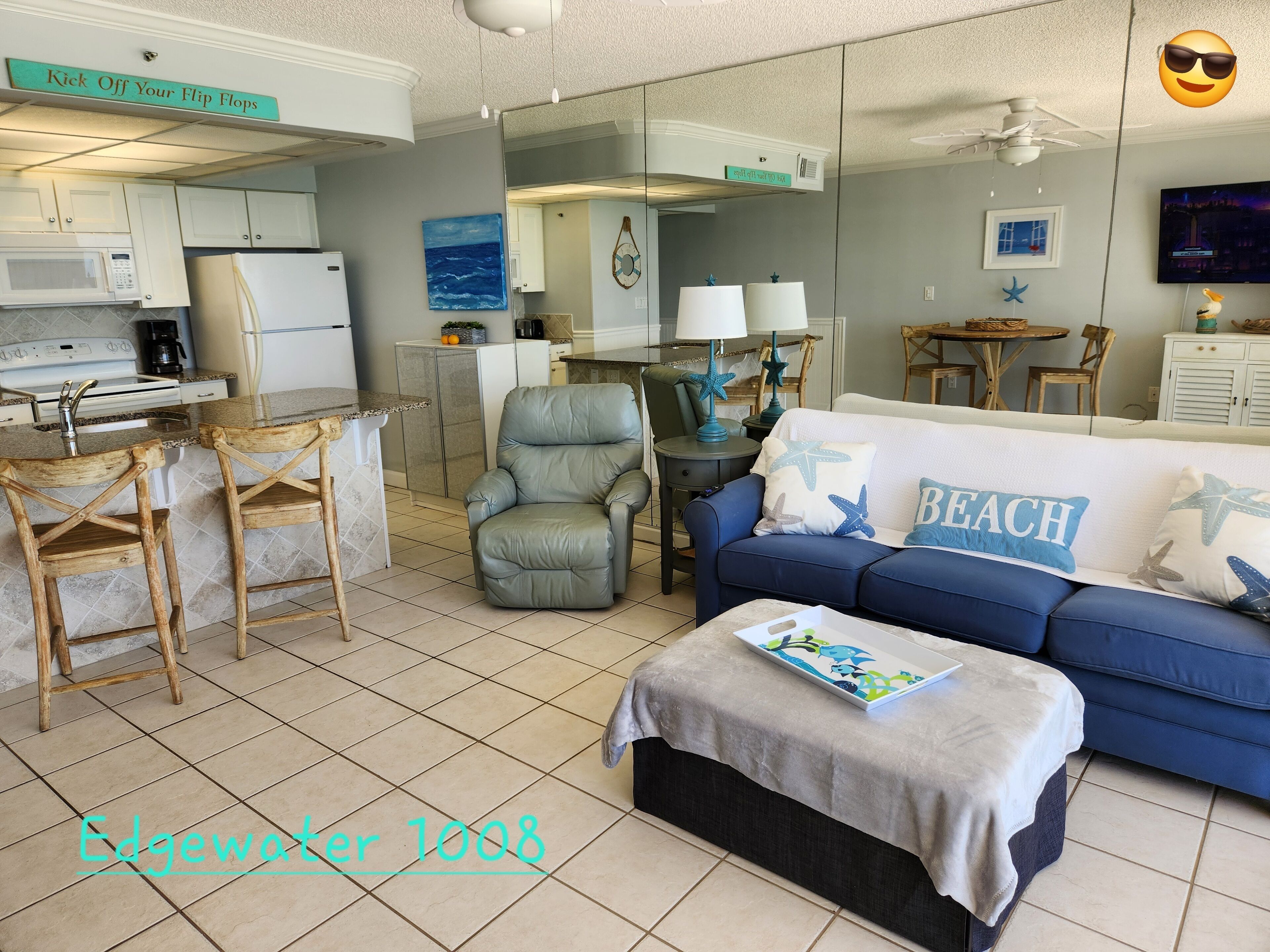Un espacio habitable compacto con kitchenette, un sofá azul con almohadas que dicen 'BEACH' y un pequeño comedor.