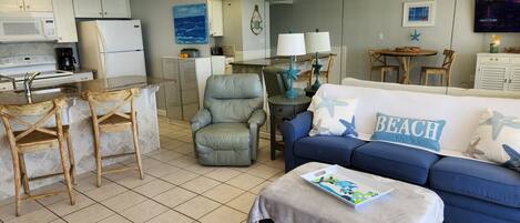 A compact living space with a kitchenette, a blue sofa with 'BEACH' pillows, and a small dining area.