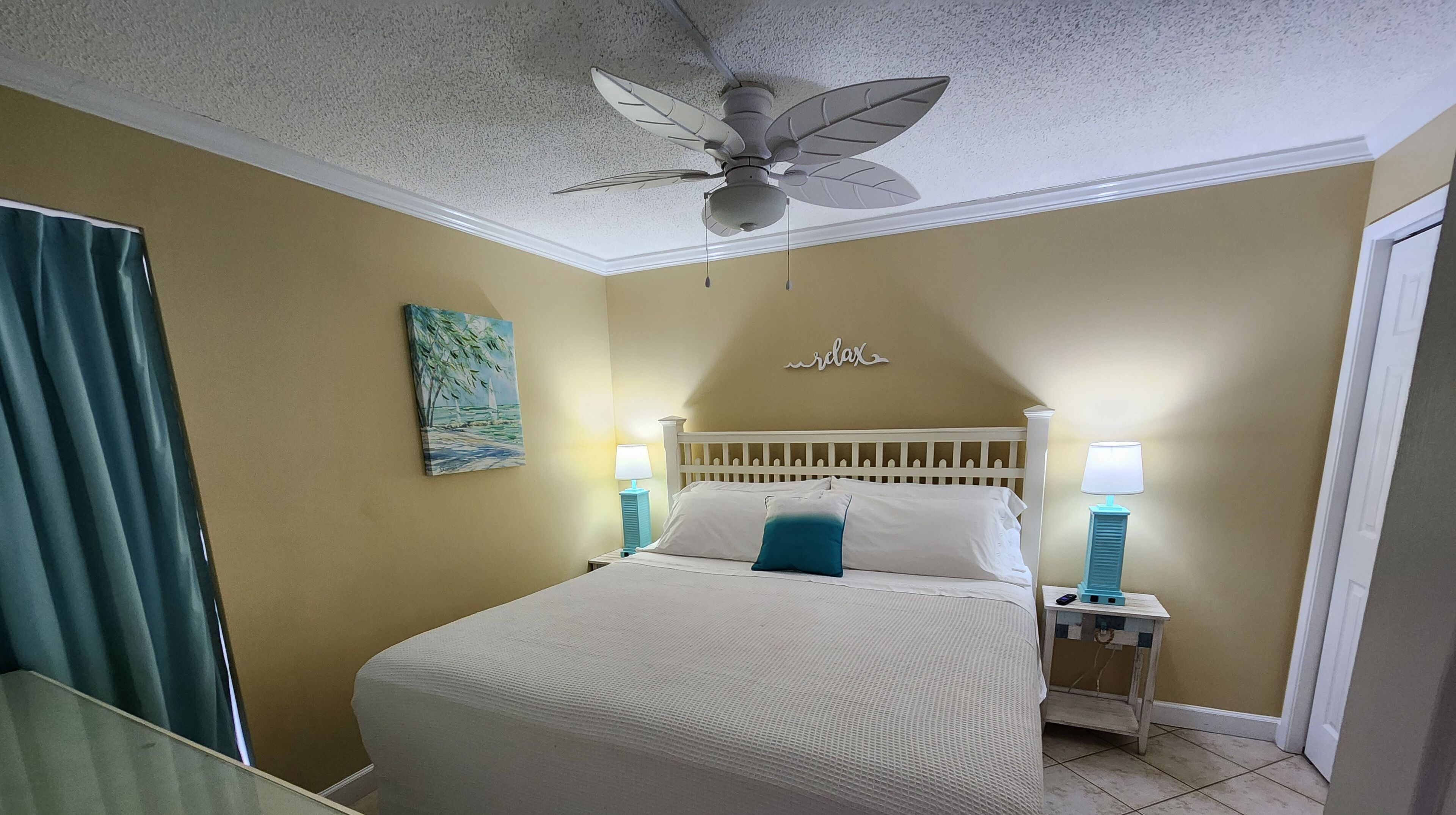 Une chambre d’hôtel avec un lit, deux tables de chevet avec des lampes, un ventilateur de plafond, un tableau au mur et une porte.